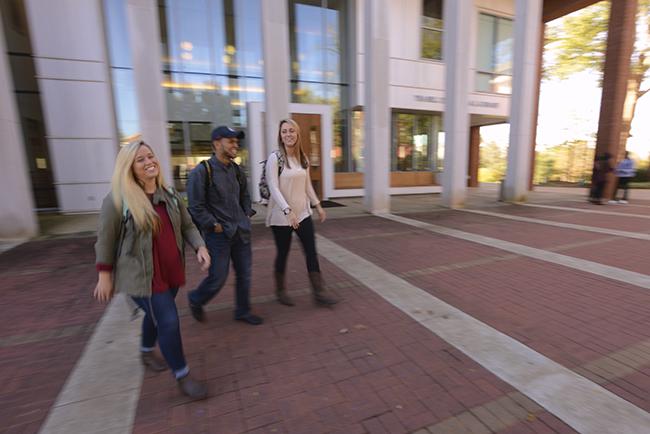 Three smiling students walk past the library on the plaza.